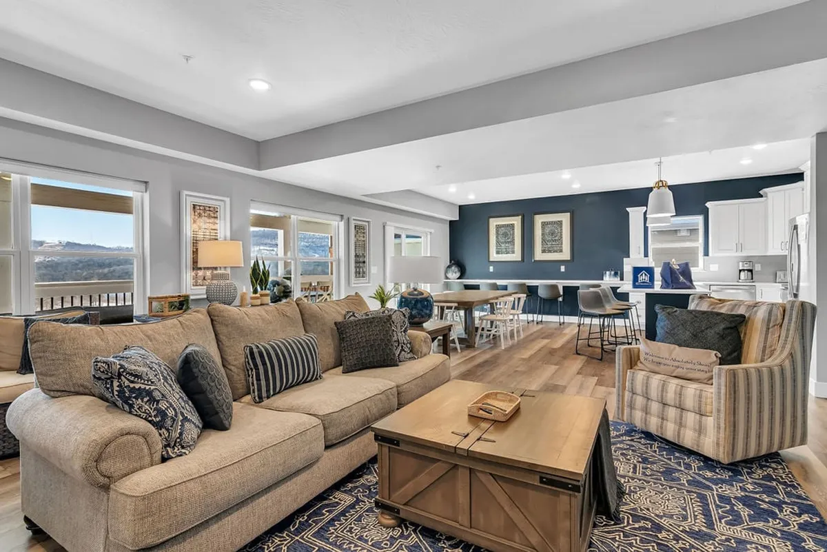 The image showcases a spacious, open-concept living area with a plush sofa, a stylish coffee table set up for gatherings, a large dining table perfect for shared meals, and a modern kitchen in the background. Sunlight pours in through expansive windows, revealing breathtaking sunset views over the lake. The scene radiates warmth, comfort, and the promise of memorable, shared moments.