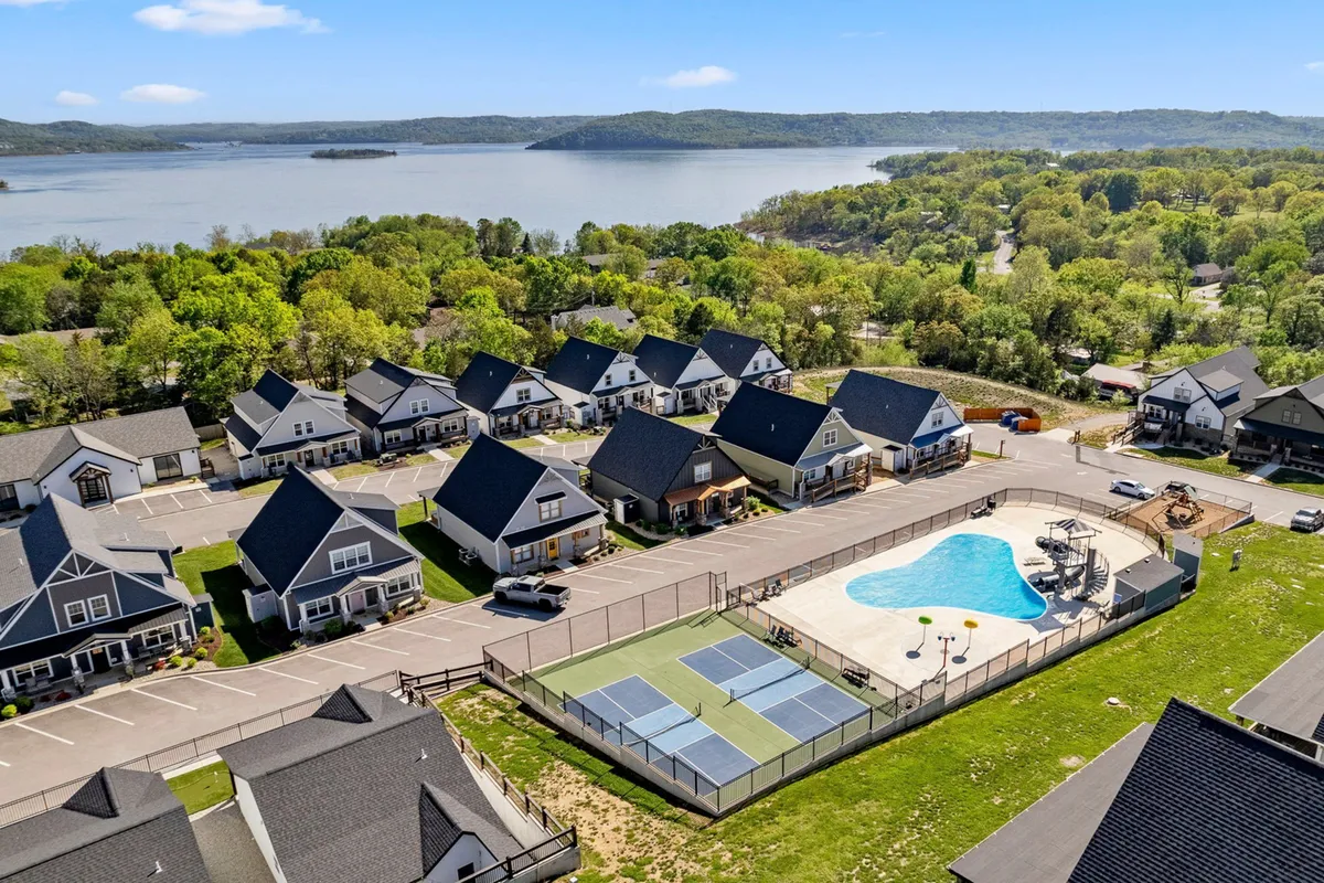 An exterior scene showing a swimming pool, tennis courts, houses, and a scenic lake view in the background. The setting is vibrant and active, hinting at a resort-style experience and social community amenities.