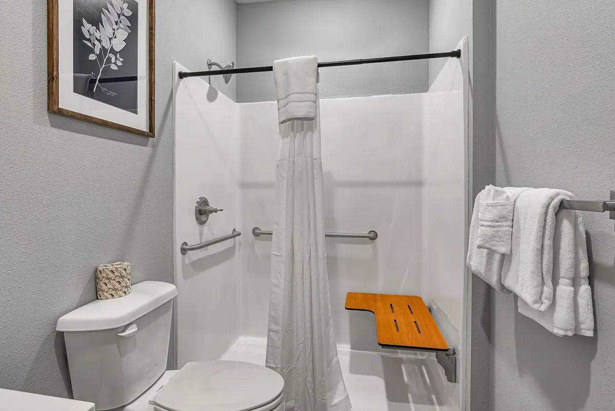 An elegant bathroom with a spacious shower area, a wooden bench for accessibility, tasteful framed artwork, and fresh towels. The mood is serene and refreshing, emphasizing comfort and inclusivity.