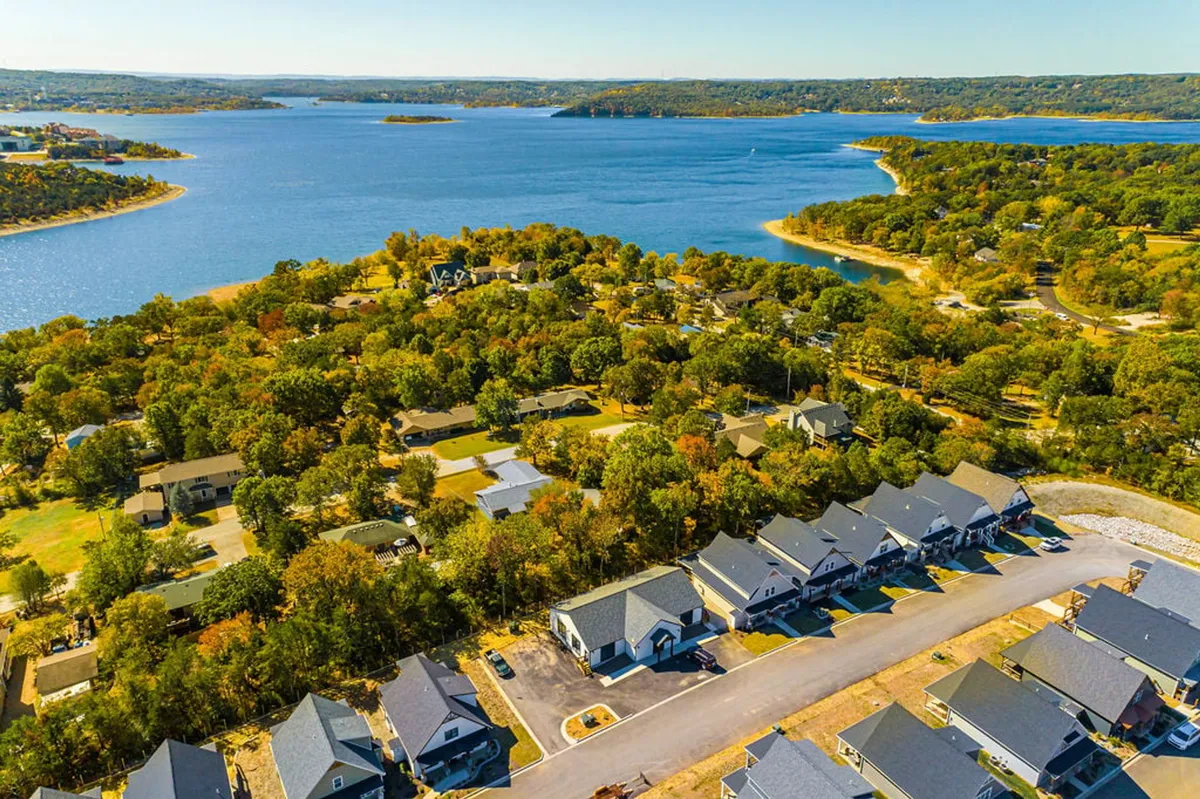 A peaceful lakeside setting showing Table Rock Lake bordered by trees with homes in the background and a road winding along the shore. The image evokes a sense of tranquility and highlights natural beauty and outdoor accessibility.