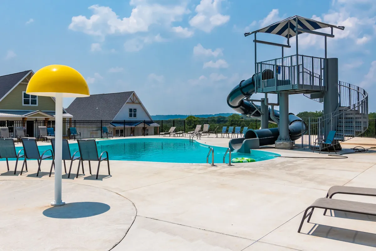 This image captures a vibrant outdoor swimming pool with a water slide, colorful lounge chairs, a yellow umbrella, and nearby houses. Children can be seen playing on the slide, while others enjoy the pool, all set against a backdrop of the community's homes.