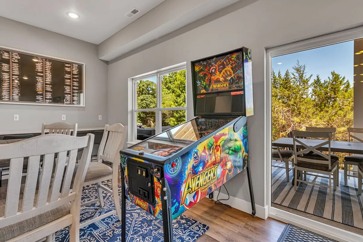 A welcoming lower level room featuring an Avengers pinball machine, a dining table, and large windows letting in natural light. The dual-purpose space highlights the pinball machine and the potential for both dining and socializing.