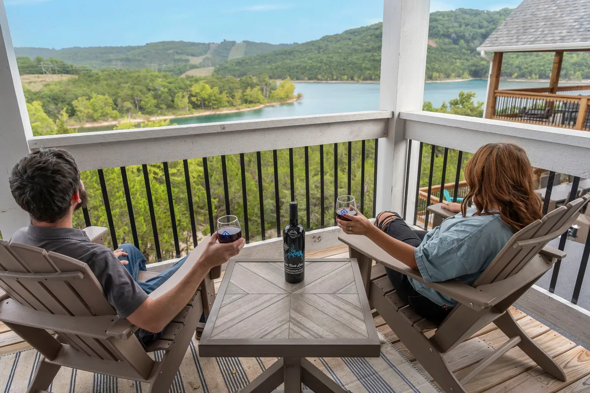 A couple sits together on outdoor chairs with a bottle of wine on the table between them. The scenic background features a beautiful lake and rolling hills at sunset, creating a tranquil, celebratory atmosphere.