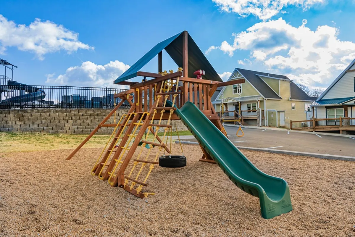 The photo showcases a well-equipped playground featuring a large play structure with a slide and swing set, surrounded by green grass. Children are captured in mid-play, enjoying the outdoors with families nearby.
